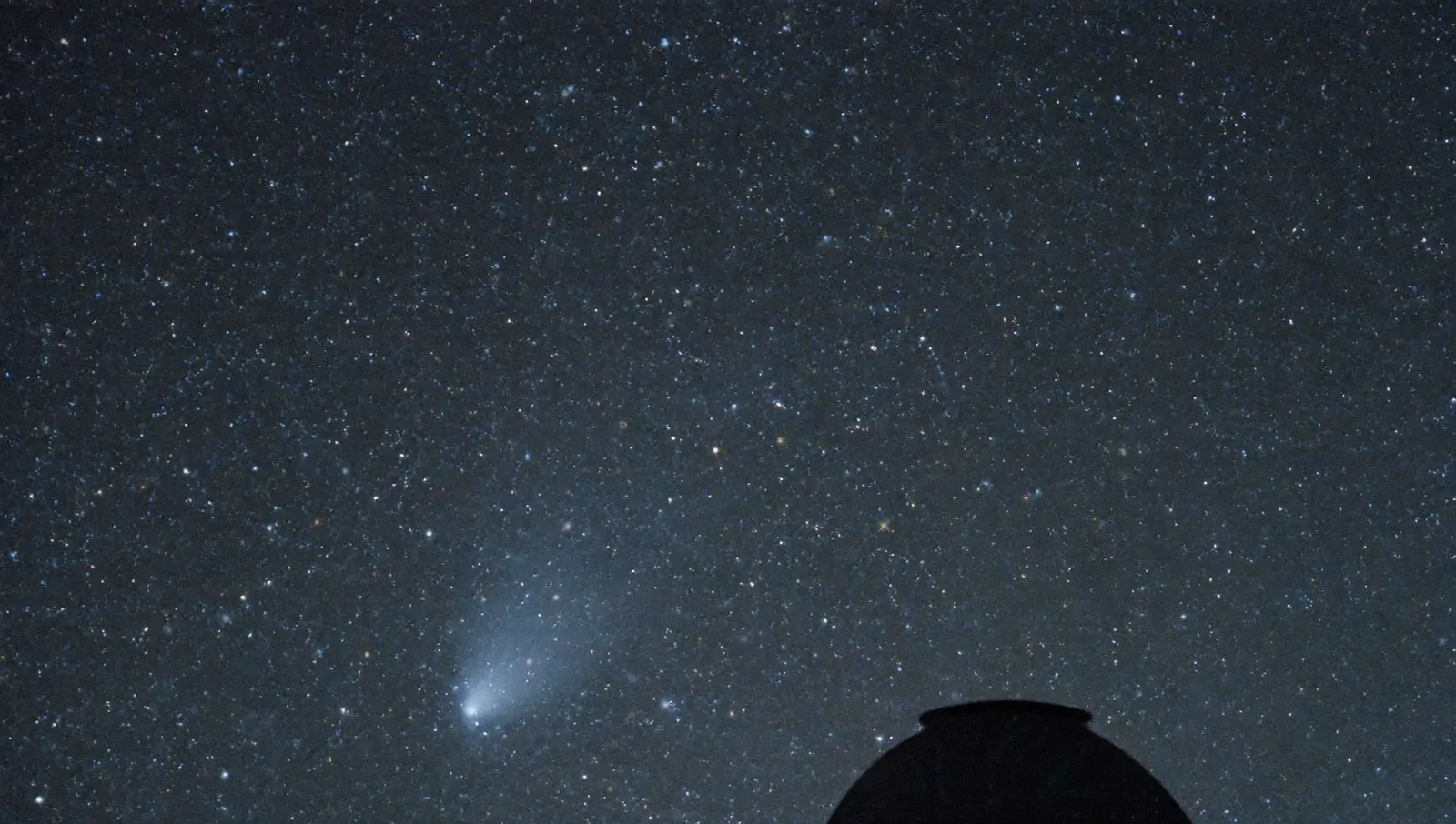 Long-exposure night sky photograph of a dense star field with a faint diffuse comet smudge low in the frame