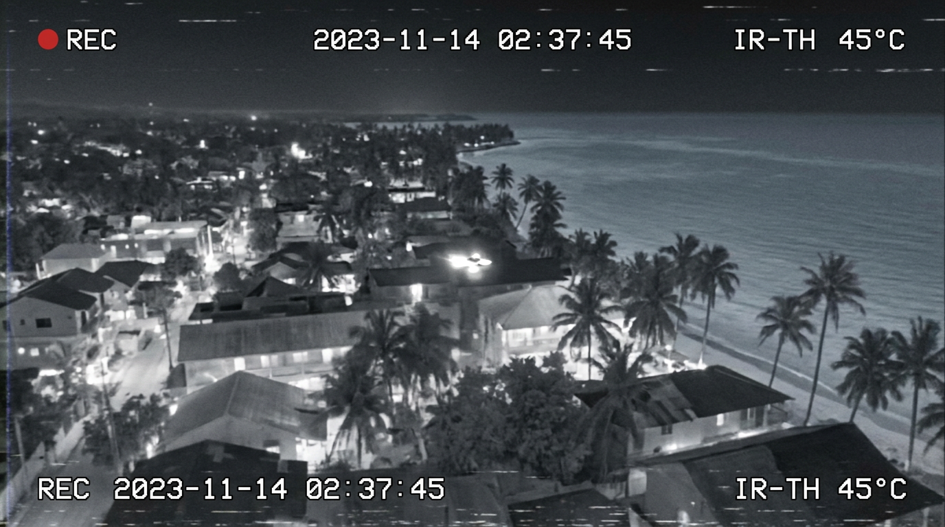 Thermal-infrared aerial view of a small bright object flying low over a tropical town's rooftops toward the coastline at night