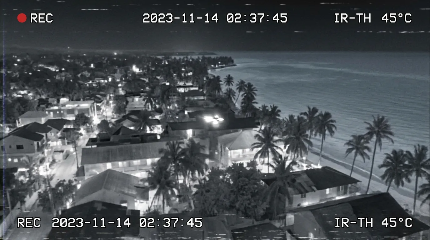 Thermal-infrared aerial view of a small bright object flying low over a tropical town's rooftops toward the coastline at night