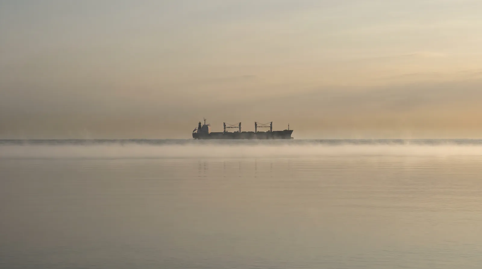 A sharp flat layer of mist lying just above a dead-calm sea at dawn, with a distant ship stretched and distorted by mirage above the layer