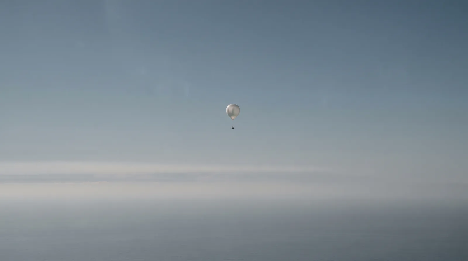 A small pale weather balloon drifting alone in a vast washed-out sky high above open ocean