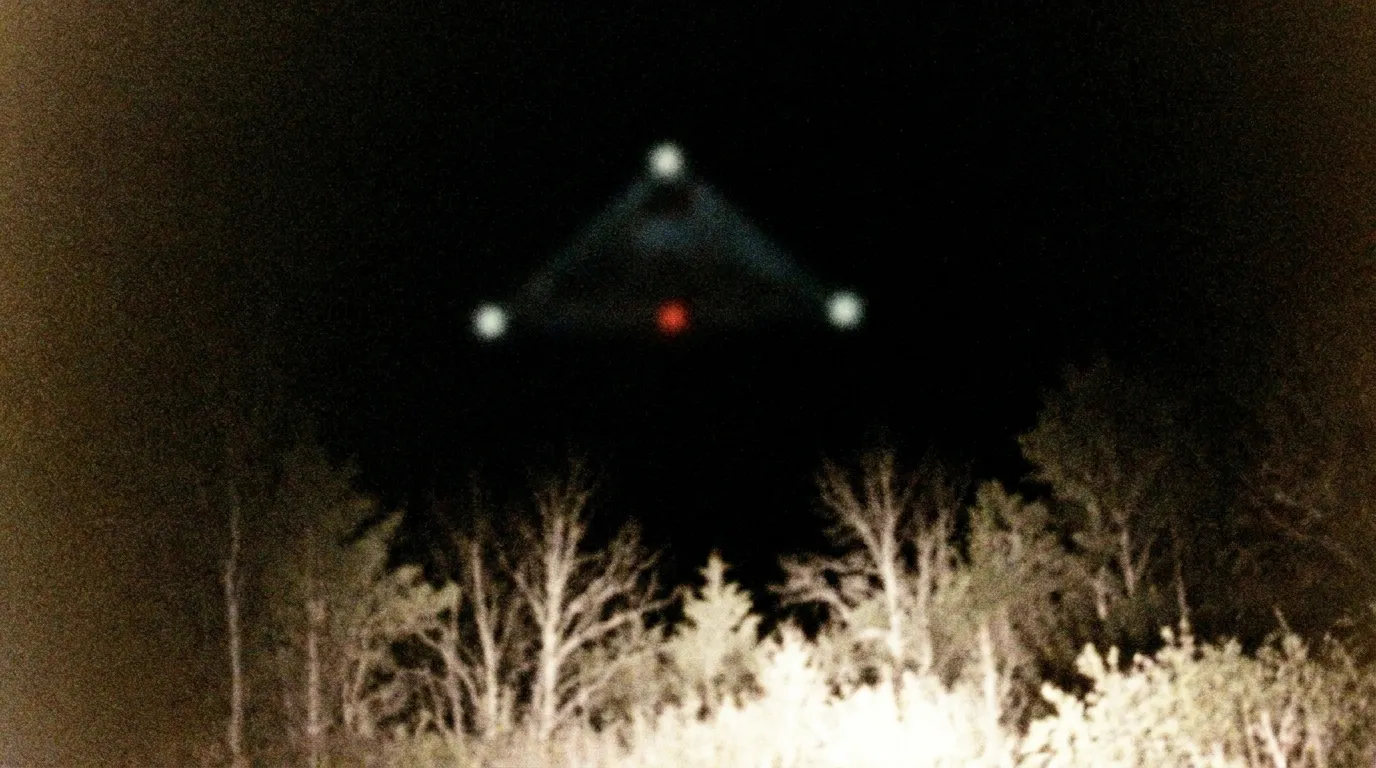 A grainy, blurry night photograph of a black triangle with three white corner lights and a central red light