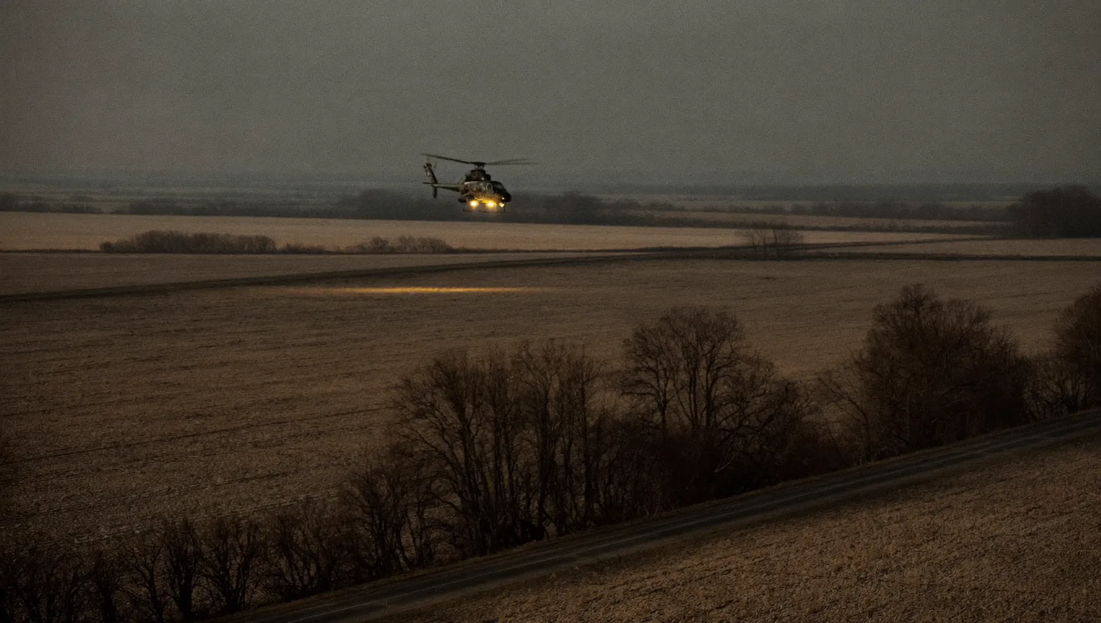 A helicopter flying low at night over flat farmland with landing lights and navigation lights on, seen from a country road