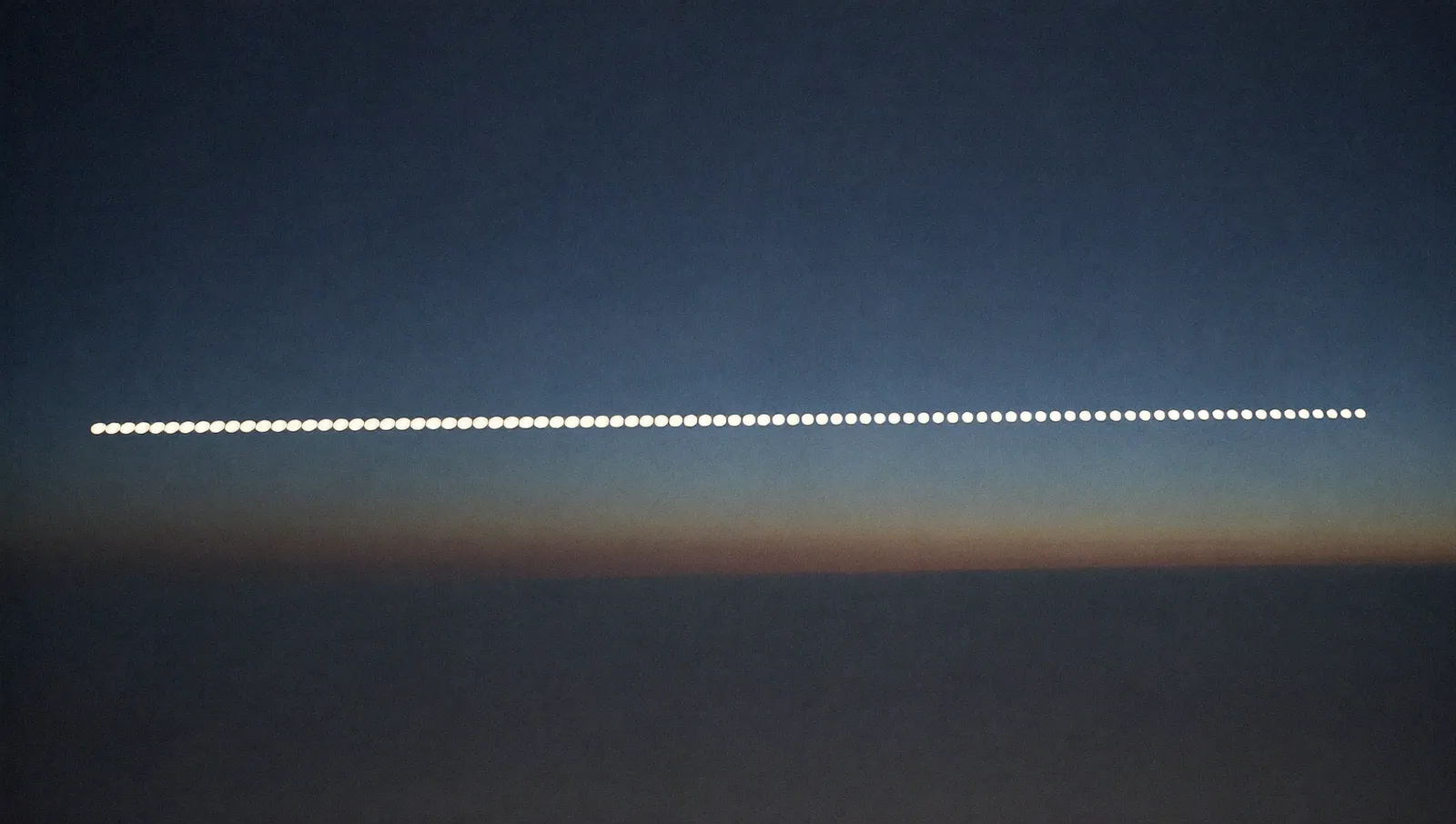 A long evenly spaced chain of bright points of light strung across a twilight sky above a layer of cloud, seen from high altitude