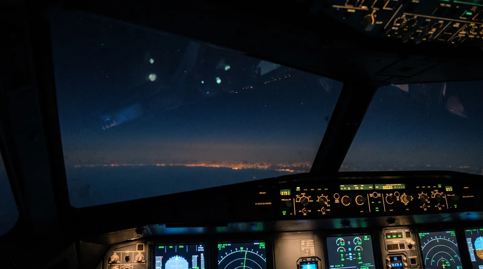 Night view from an airliner cockpit at cruising altitude, a deep indigo sky ahead and faint distant lights on the horizon far below