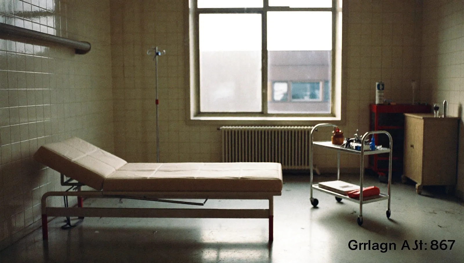 An empty 1960s hospital examination room with a made bed, a stainless steel instrument trolley and flat daylight from a tall window