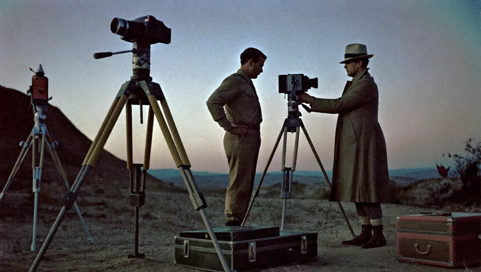 Period military officers and a civilian scientist in overcoat and fedora standing at tripod-mounted cameras and a theodolite in the New Mexico desert at dusk