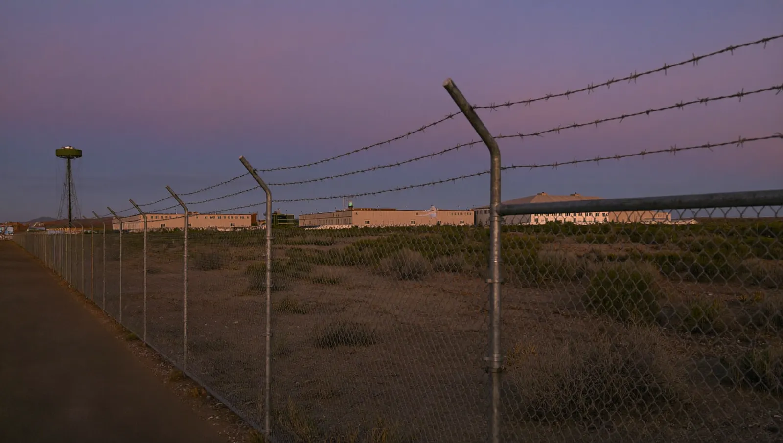 A chain-link and barbed-wire perimeter fence crossing high desert scrub at dusk, with low government buildings and a water tower beyond