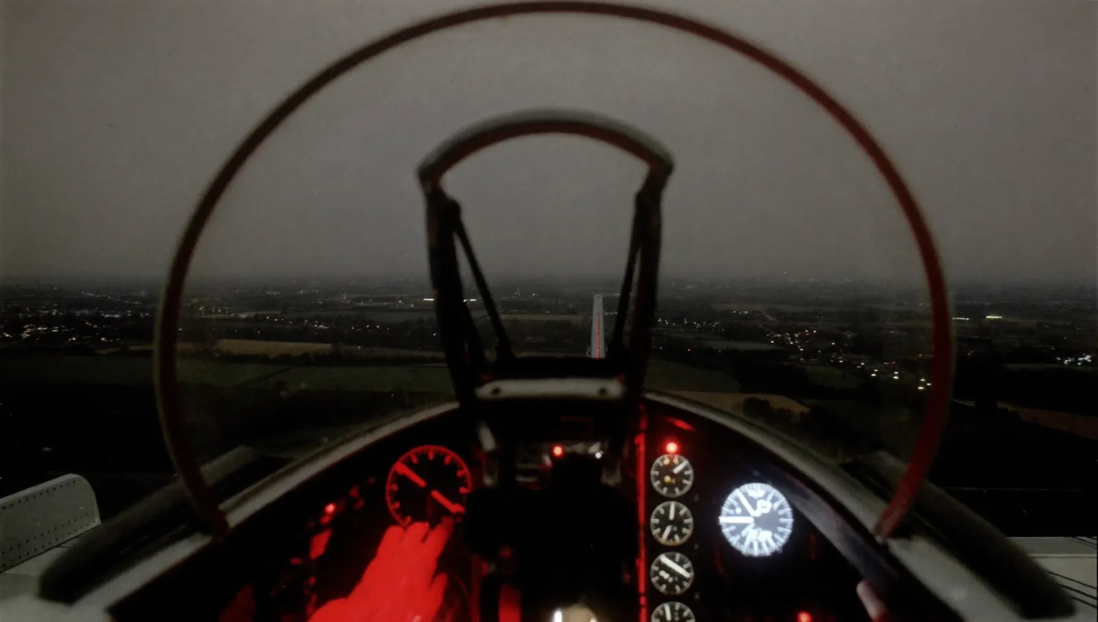 View forward from a 1950s jet night-fighter cockpit at night over flat dark farmland with scattered village lights