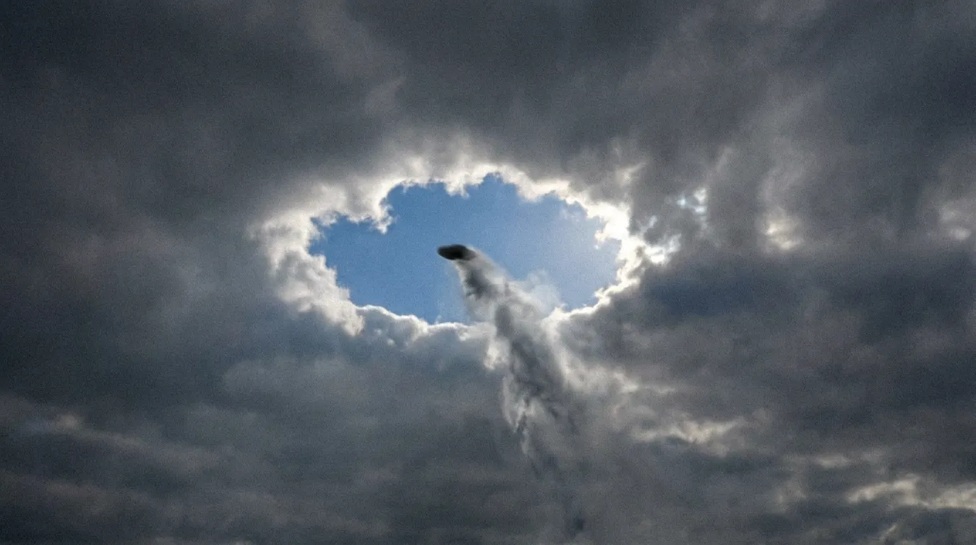 A dark disc shooting upward punching a clean circular hole through a grey overcast cloud layer