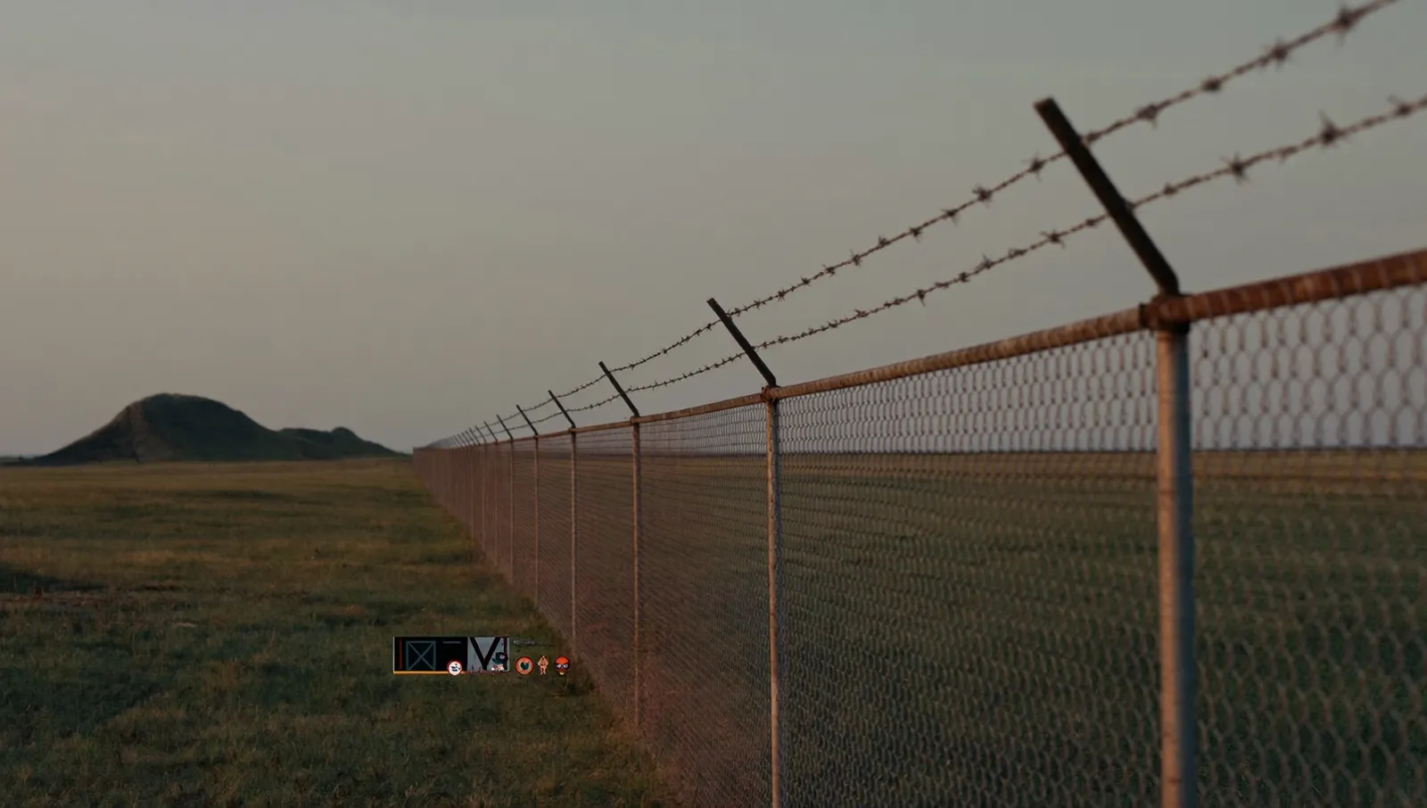 A chain-link security fence topped with razor wire running across flat Texas Panhandle grassland at dawn, with low bunker mounds in the distance