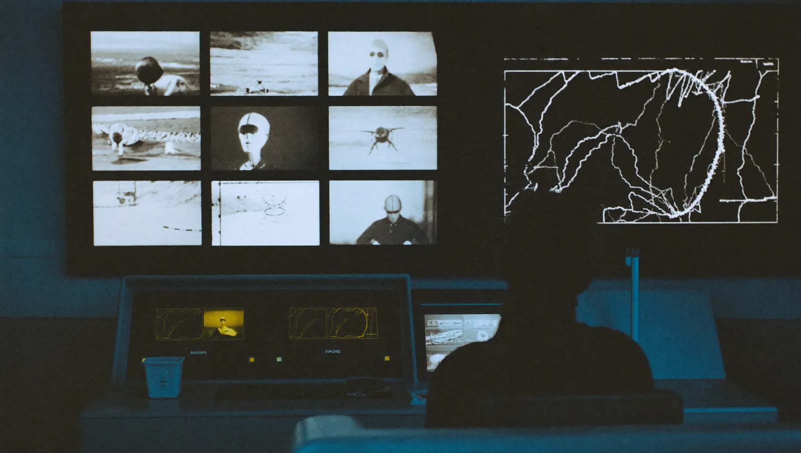 A security control room with a wall of monochrome camera feeds and a radar display, a single operator silhouetted at the console