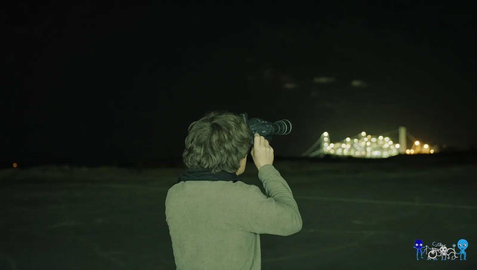 A lone figure seen from behind on dark open ground at night holding a night-vision monocular to one eye, a floodlit facility on the horizon