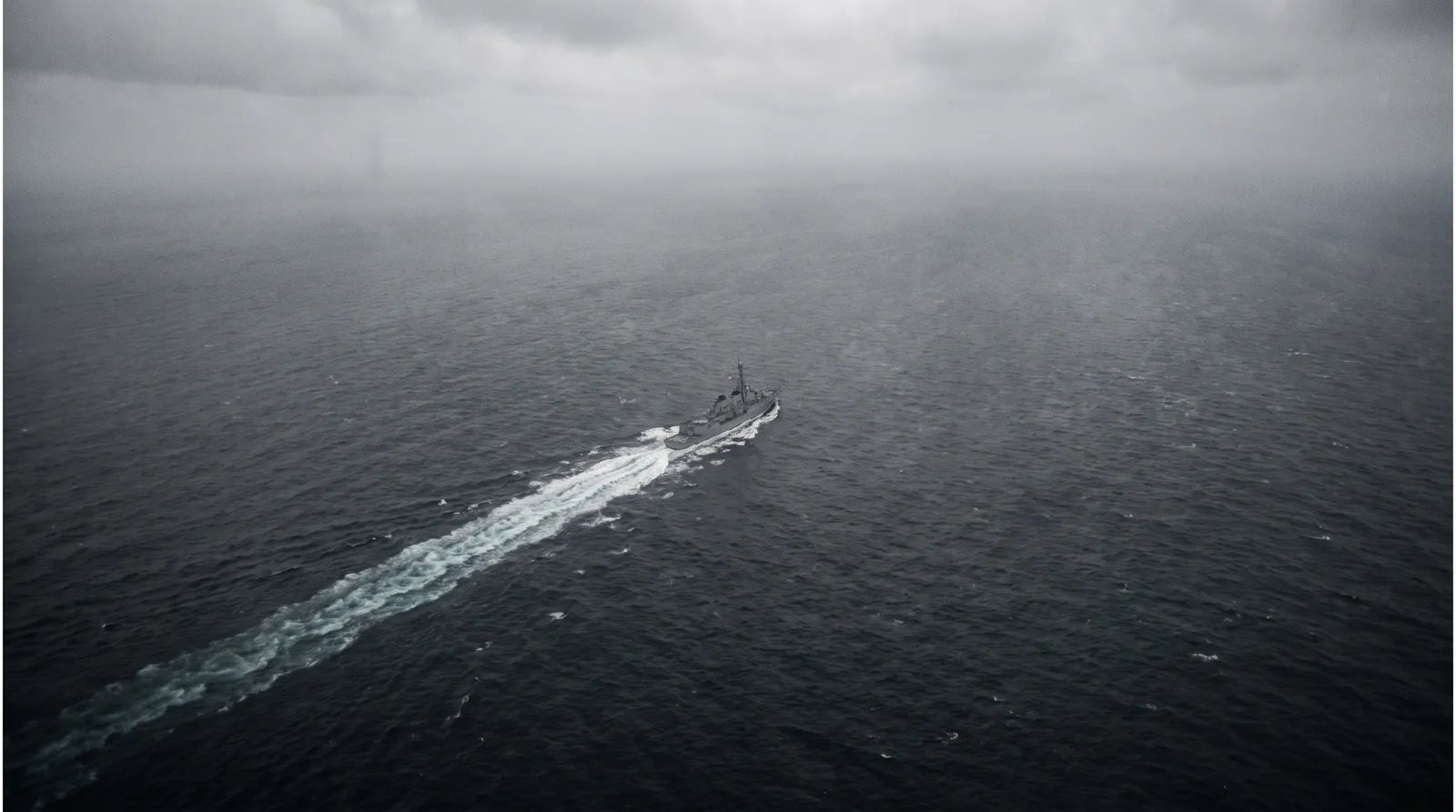 High-altitude aerial view looking down at a grey warship under way on dark open ocean with a long white wake behind it