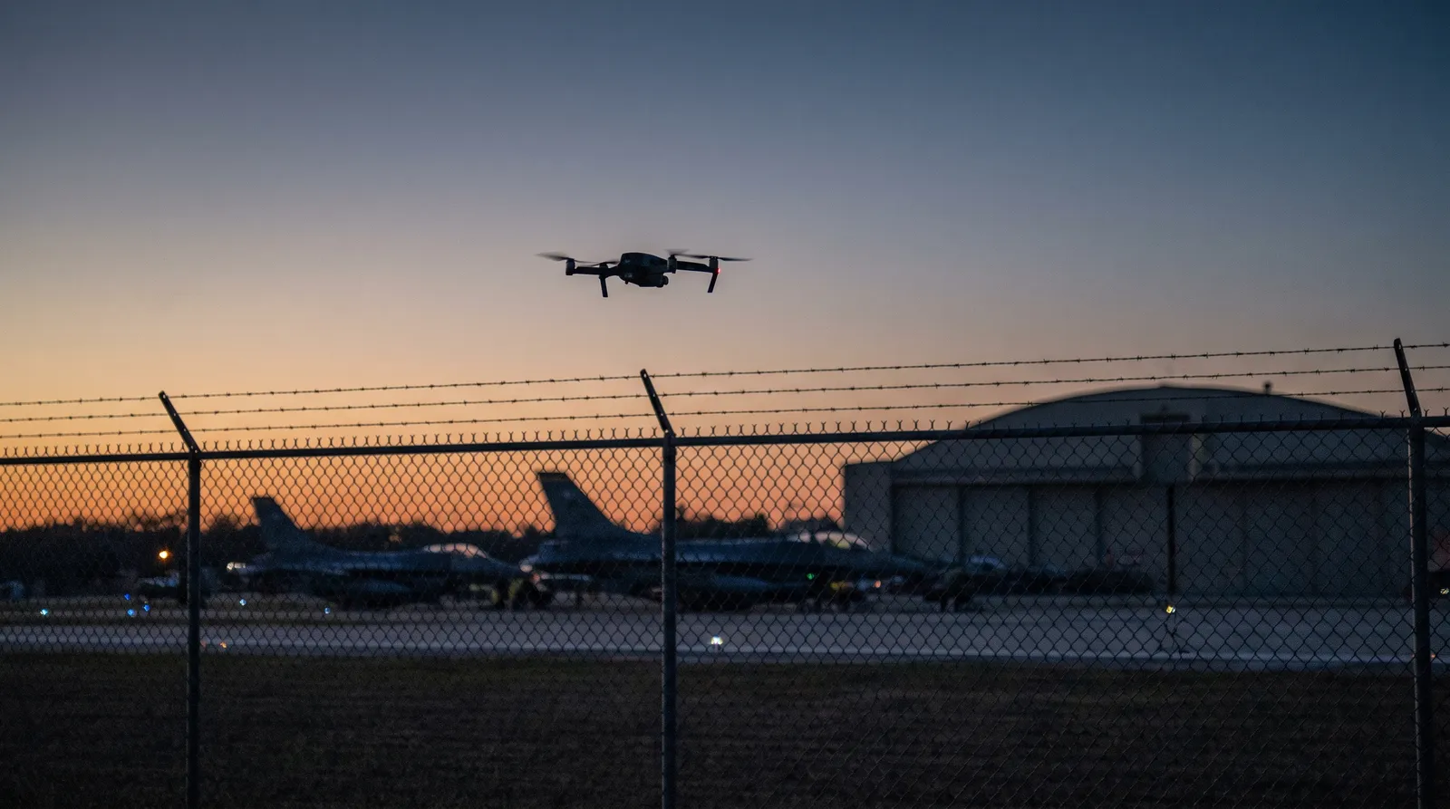 A small dark quadcopter drone silhouetted at dusk above a military airfield perimeter, with parked fighter jets blurred behind it