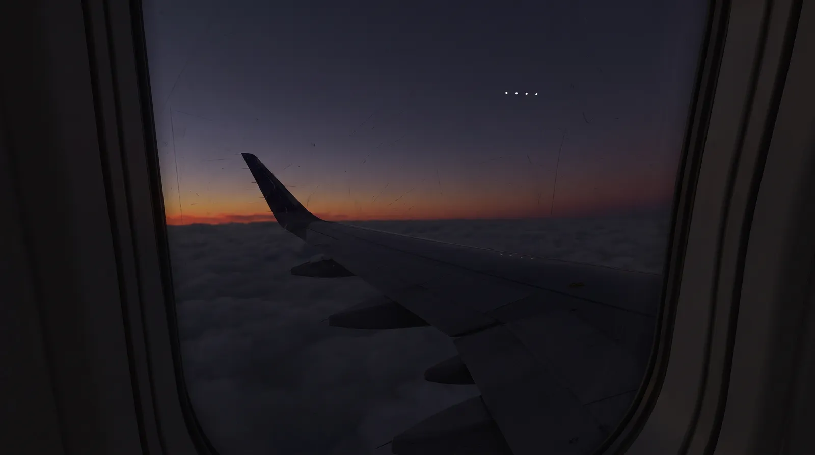 The view from an airliner window at deep twilight: a flat deck of cloud below, a band of orange fading to indigo, and four bright points of light high above the wing