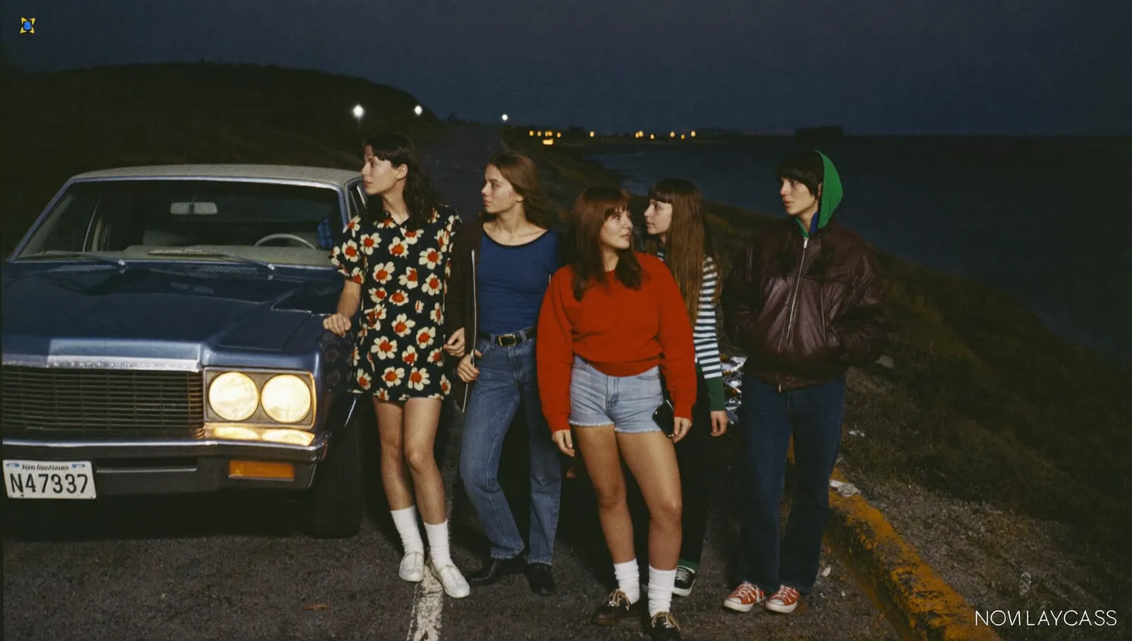 Five teenagers in 1967 clothing standing beside a parked car on a dark Nova Scotia coastal highway at night looking at distant lights over black water
