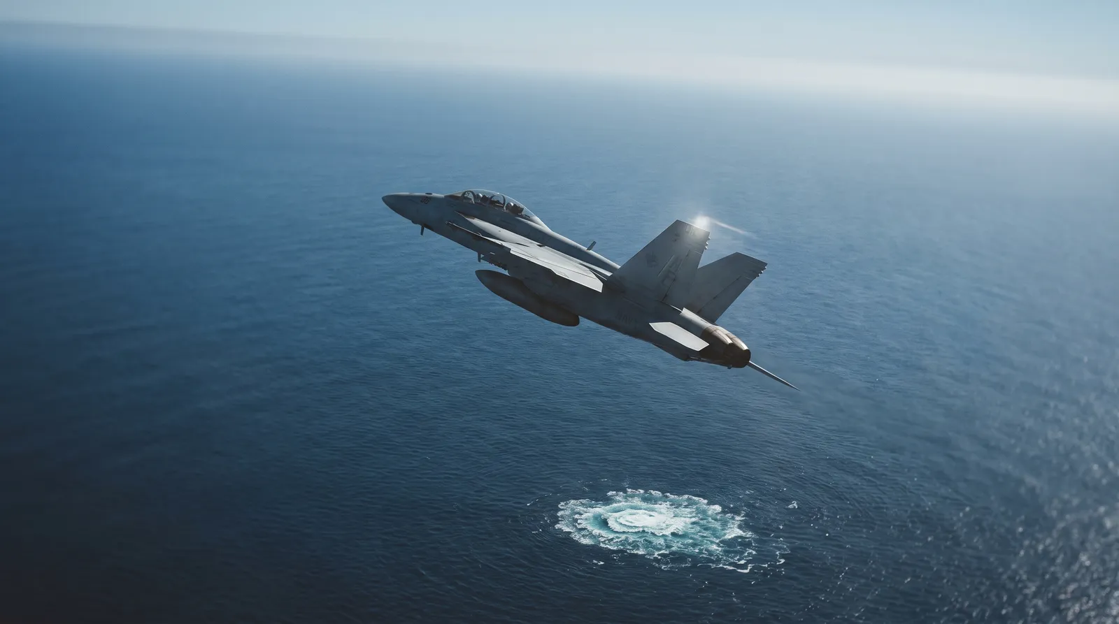 A US Navy F/A-18F Super Hornet banking over the open Pacific in bright daylight, a patch of disturbed white water on the sea below
