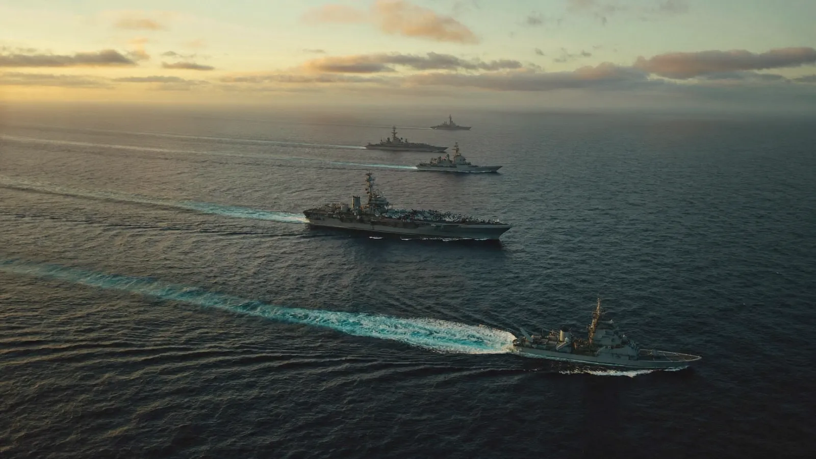 Aerial view of a US Navy carrier strike group underway at dawn, an aircraft carrier and a guided-missile cruiser trailing long wakes on a calm Pacific