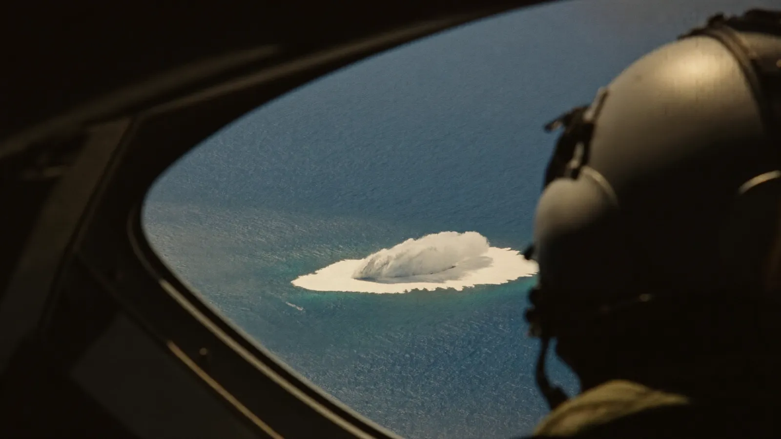 View over a fighter pilot's shoulder from an F/A-18 cockpit, looking far down at an oval patch of churning white water on a calm deep blue ocean