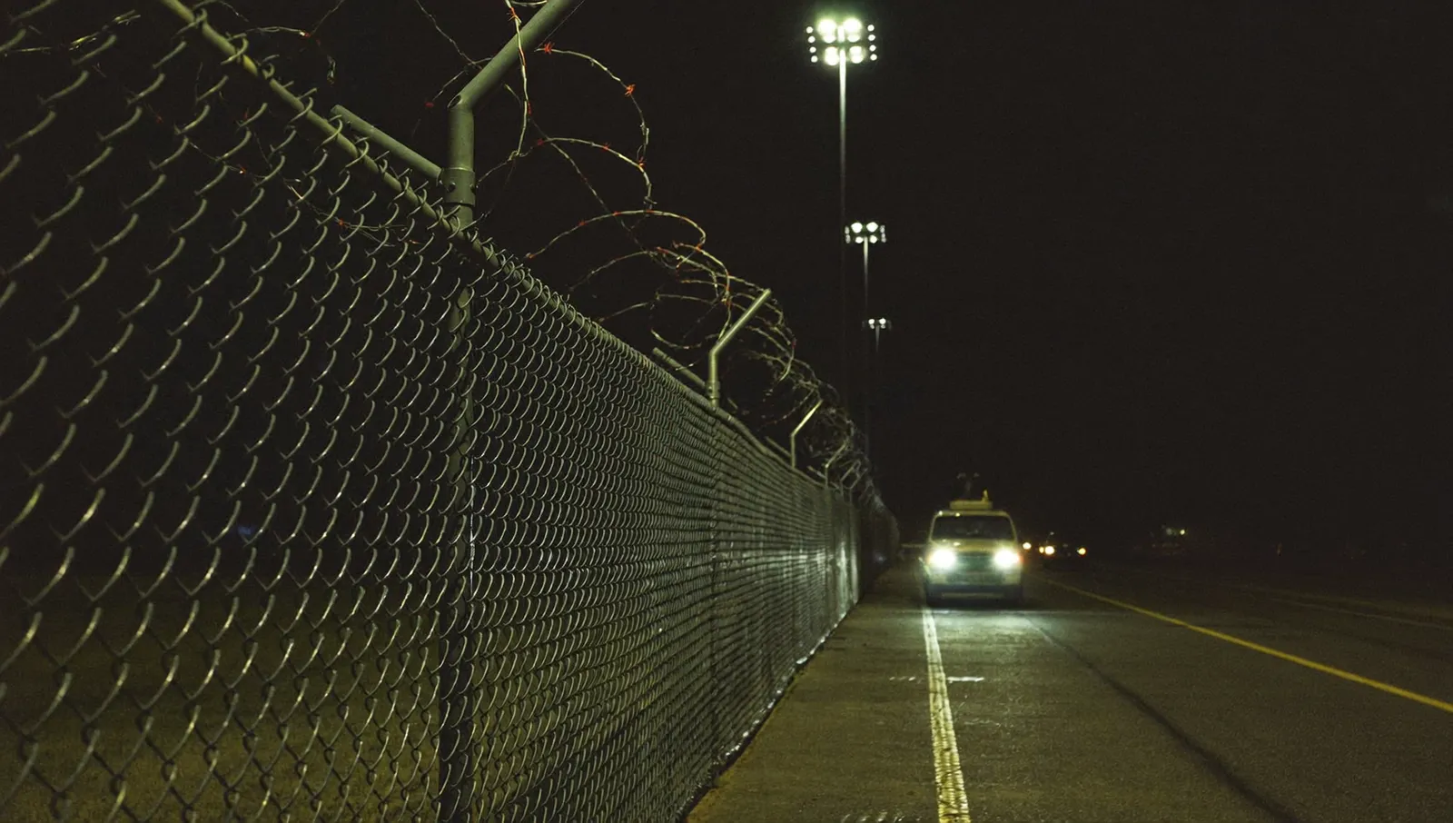 A security patrol vehicle with headlights on beside a razor-wire perimeter fence at a military airbase at night
