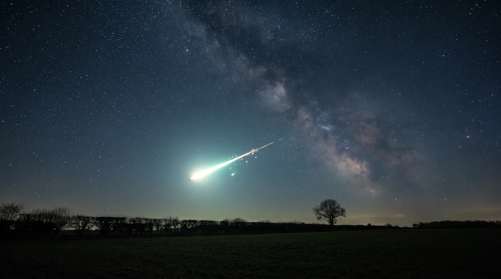 A brilliant meteor fireball streaking low across a dark night sky above open countryside, leaving a glowing trail