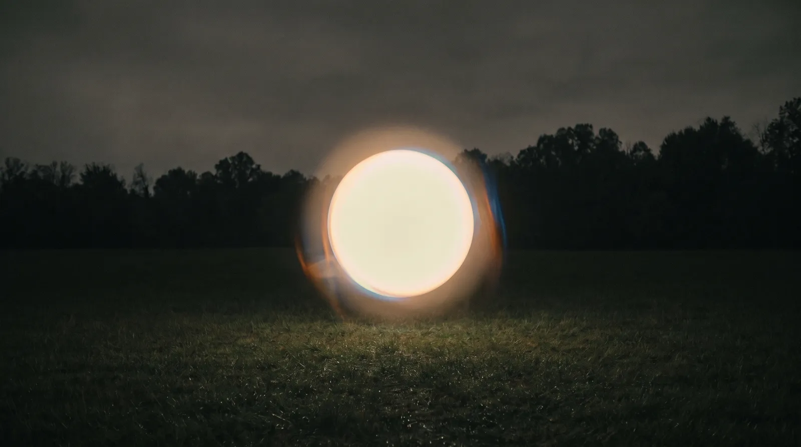 A single glowing pale sphere hovering low over a dark empty field at night, ringed by a soft fuzzy corona of light
