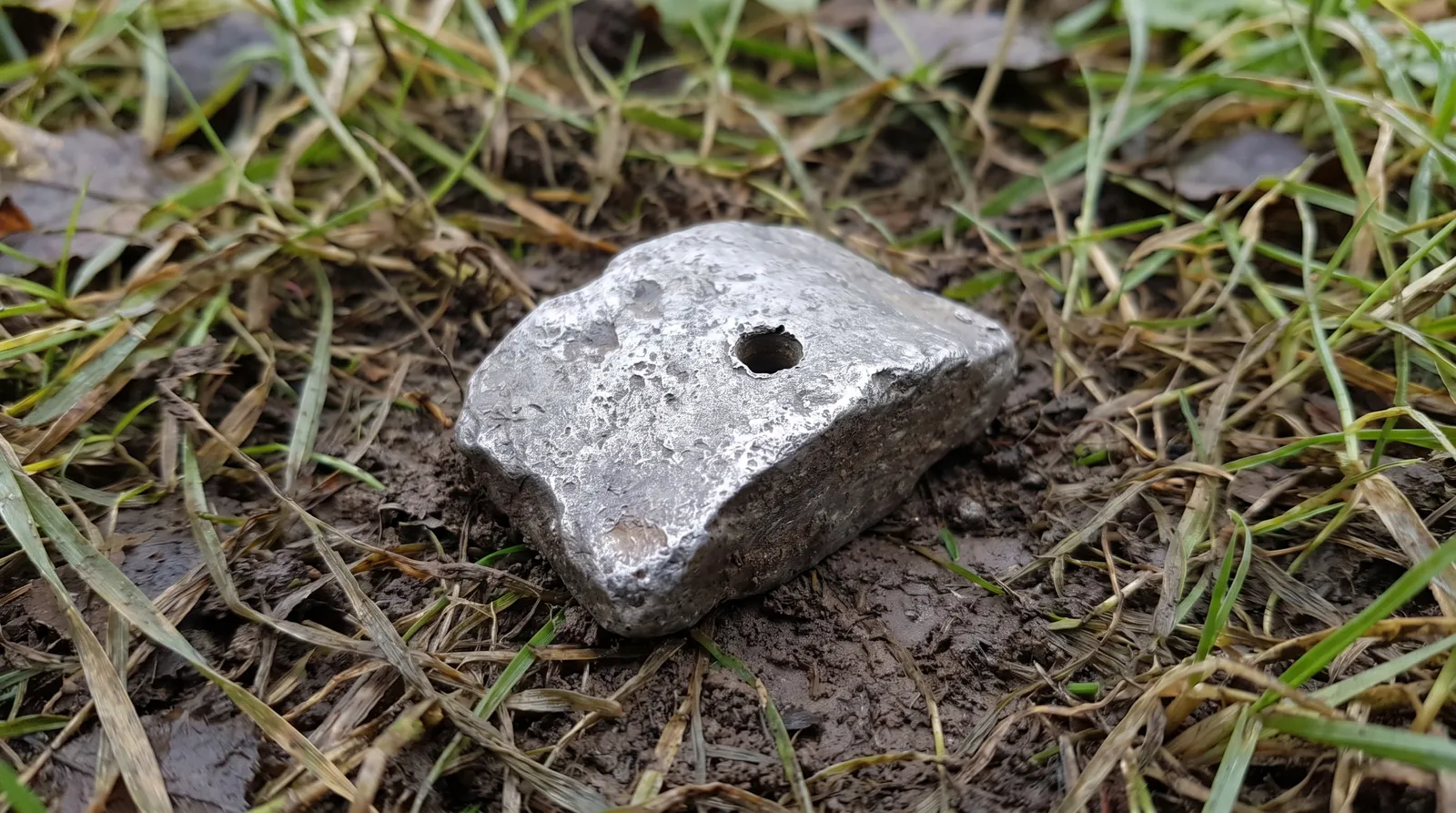 A small irregular lump of dull grey cast metal with a drilled hole, lying half-embedded in damp dark soil and flattened grass