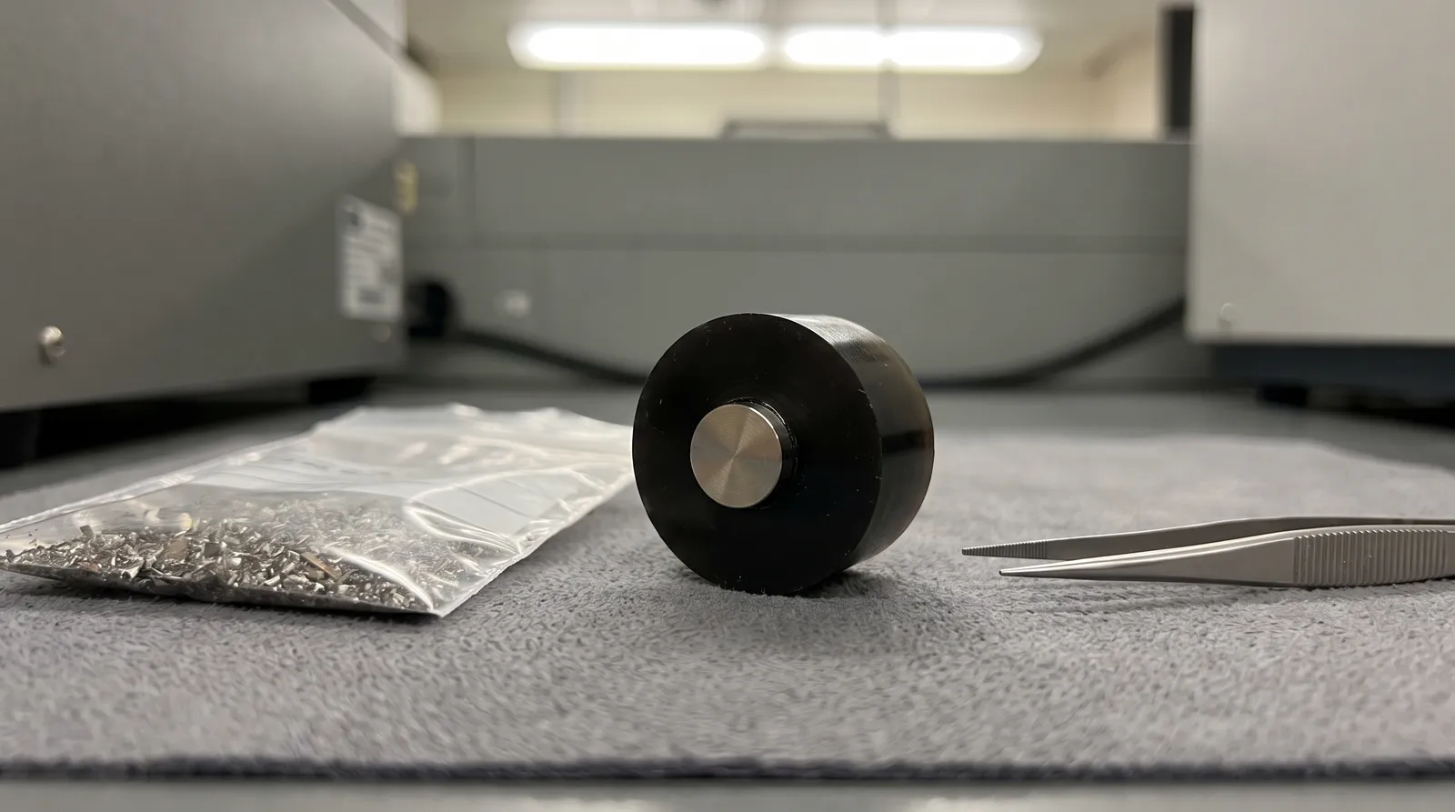 A polished metal sample mounted flush in a black resin puck on a laboratory bench beside steel tweezers and a sealed bag of metal shavings