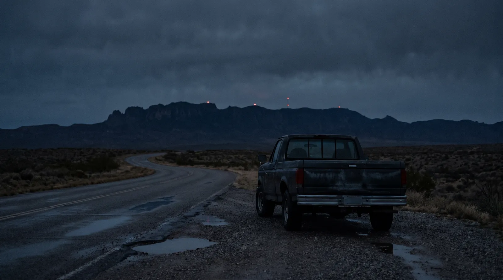 A pickup truck parked on the gravel shoulder of an empty desert road at night, with small red lights hanging above a distant mountain ridge