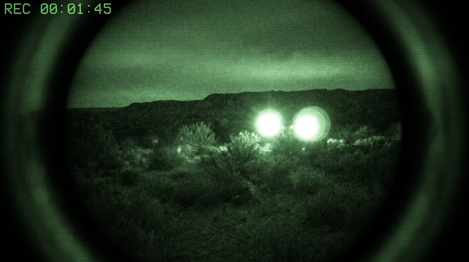 The view through a night-vision image intensifier: a grainy green monochrome landscape with two blooming white points of light flaring into soft discs
