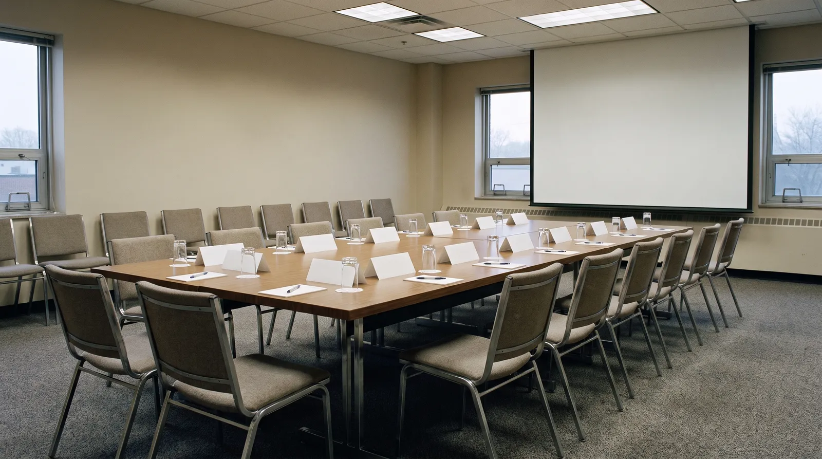 A plain institutional conference room set for a morning meeting, a long table ringed with chairs, blank name cards and notepads at each place